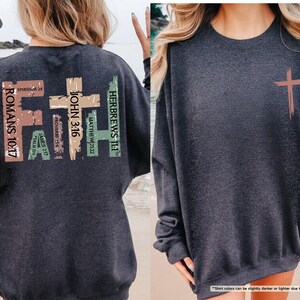 May include: Dark heather grey sweatshirt with a large graphic on the back. The graphic features a cross with the word "FAITH" written vertically in different colors. The cross is surrounded by bible verses, including Romans 10:17, Hebrews 11:1, John 3:16, Matthew 21:22, Proverbs 15:6, James 2:17, and Psalm 105:4.