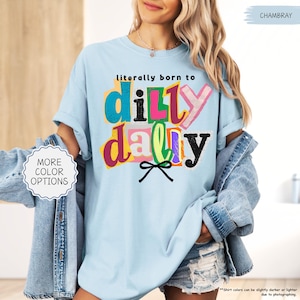 K&ouml;nnte beinhalten: Hellblaues T-Shirt mit dem Text "literally born to dilly dally" in bunten, geschichteten Buchstaben. Das Shirt hat eine lockere Passform und kurze &Auml;rmel. Eine Jeansjacke wird &uuml;ber dem Shirt getragen.