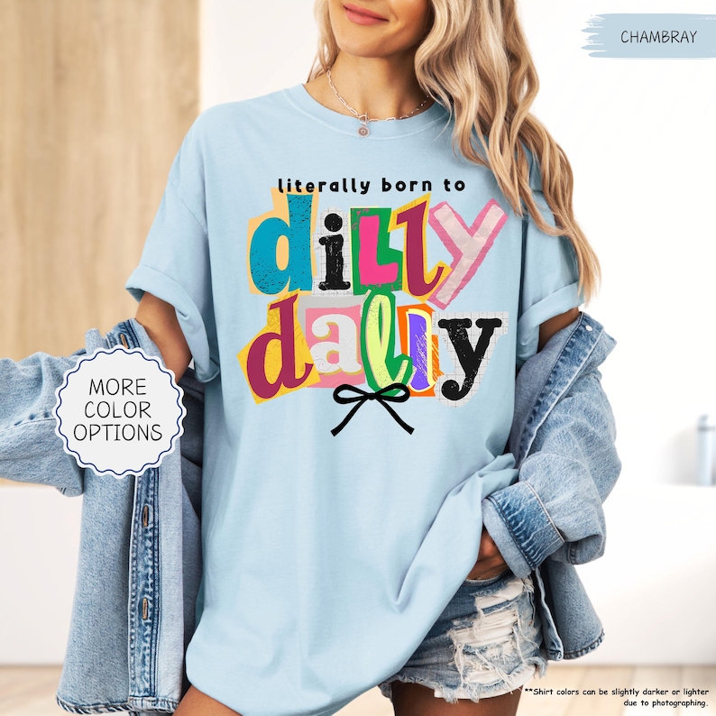 K&ouml;nnte beinhalten: Hellblaues T-Shirt mit dem Text "literally born to dilly dally" in bunten, geschichteten Buchstaben. Das Shirt hat eine lockere Passform und kurze &Auml;rmel. Eine Jeansjacke wird &uuml;ber dem Shirt getragen.
