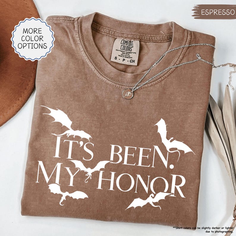 Pu&ograve; includere: T-shirt color espresso con la scritta bianca "IT'S BEEN. MY HONOR" e grafica di pipistrelli bianchi. La maglietta ha girocollo e maniche corte. Una collana d'argento con un ciondolo &egrave; drappeggiata sulla maglietta. Marchio Comfort Colors.