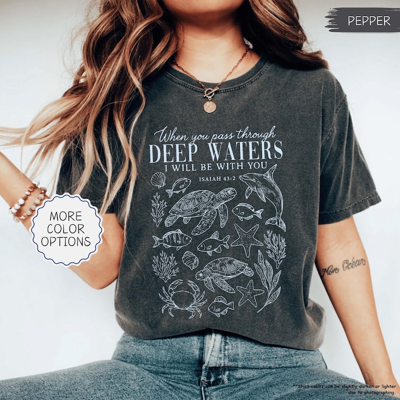 May include: Pepper-colored t-shirt with a white graphic featuring marine life and the text "When you pass through deep waters, I will be with you, Isaiah 43:2." The shirt has a crew neck and short sleeves. "More color options" is also displayed.