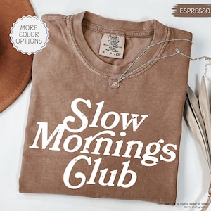 May include: A brown t-shirt with the words "Slow Mornings Club" printed in white. The shirt has a crew neck and short sleeves. A silver necklace with a small pendant is draped across the shirt. The words "More Color Options" are in a white circle.