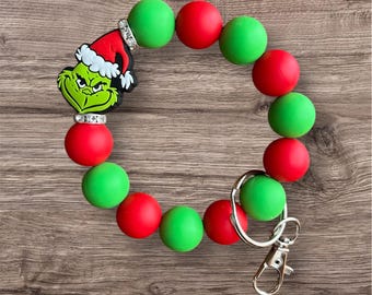 May include: A green and red beaded bracelet with a Grinch character charm and a silver key chain. The bracelet is made of silicone beads.