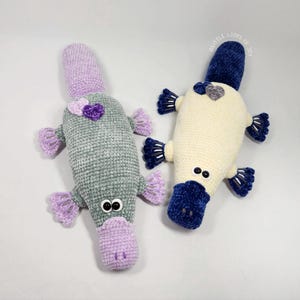 May include: Two handmade crochet platypus toys. One is gray with purple accents and a purple heart. The other is cream-colored with navy blue accents. Both have button eyes and are lying on a white surface.