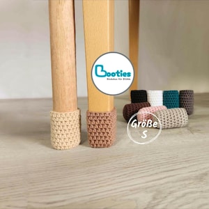 Könnte beinhalten: Kleine gehäkelte Stuhlbeinüberzieher in verschiedenen Unifarben wie Beige, Braun, Schwarz, Weiß, Türkis und Rosa. Zwei Überzieher stecken an hellen Holzstuhlbeinen, weitere liegen in einer Gruppe daneben. Der Text lautet Booties und Größe S.