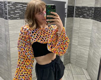 May include: A person wearing a colorful crocheted crop top with long sleeves. The top is made of a variety of colors, including yellow, orange, pink, and blue. The person is wearing black shorts and has their hair pulled back.