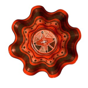 May include: A decorative, wavy-edged bowl with a vibrant, layered design. The bowl features alternating bands of orange and brown, with a central starburst design in shades of orange, brown, and beige. Small black dots accent the design.
