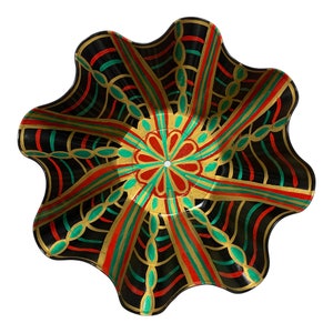 May include: A decorative black bowl with a scalloped edge. The bowl features a colorful geometric design with red, green, and gold accents. The design radiates from the center, creating a starburst effect. The bowl is likely used for serving or display.