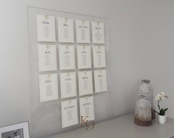 Könnte beinhalten: Transparente Tischordnung aus Acryl mit 15 weißen Karten, die jeweils mit einer transparenten Klammer befestigt sind. Die Karten haben schwarzen Text und sind in einem Raster angeordnet. Unten befindet sich ein goldenes Monogramm. Dekorative Vasen und eine Topfpflanze.