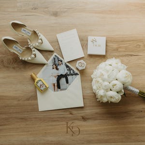May include: A flat lay of wedding accessories on a wooden surface. Features white Jimmy Choo heels with pearl detailing, a bouquet of white peonies, a Dior box, an envelope with a photo, a perfume bottle, and wedding rings.
