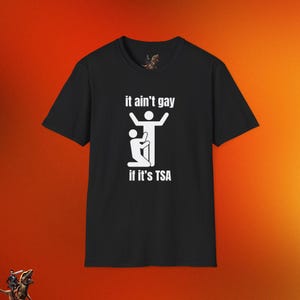 May include: Black t-shirt with white graphic text and illustration. The text reads "it ain't gay if it's TSA" above a stick figure illustration. The shirt is displayed on an orange background.