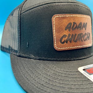 Może przedstawiać: Czarna czapka trucker z brązowym skórzanym naszywką z napisem "Adam Church" białymi literami.