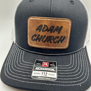 Peut inclure: Casquette de camionneur noire et blanche avec un patch en cuir marron qui dit "ADAM CHURCH". La casquette a un dos en maille blanche et un devant noir avec des détails de couture blanche. La casquette a également une étiquette noire et rouge qui dit "EST. 1970 RICHARDSON ORIGINAL 112 TRUCKER ADJUSTABLE".