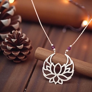 May include: Silver-toned lotus flower pendant necklace with a delicate chain. The pendant features an intricate openwork design and is accented with small, round, purple beads. The necklace is displayed on a wooden dowel, with pine cones in the background.