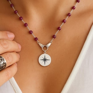 May include: A silver necklace with a pendant featuring a black compass rose design. The necklace has a chain of small, round, dark red beads. The pendant is attached to the chain with two silver hand-shaped clasps.