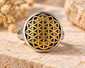 Könnte beinhalten: Ein silberfarbener Ring mit einer kreisförmigen Vorderseite, die ein goldenes, geometrisches Muster der Blume des Lebens auf dunklem Hintergrund zeigt. Der Ring hat eine polierte Metallschiene und ein flaches, rundes Design mit Linien.