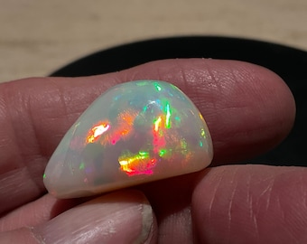 May include: A polished, oval-shaped opal gemstone. The opal has a milky white base color with vibrant flashes of red, green, and yellow iridescence. The stone is held in a hand, showcasing its smooth, rounded form and play of color.