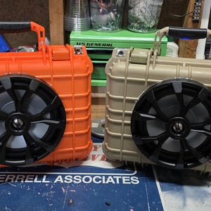 Puede incluir: Dos altavoces portátiles resistentes en la imagen, uno naranja y otro beige. Cada uno tiene una gran rejilla de altavoz negra. Están diseñados para parecerse a maletas resistentes, con un asa en la parte superior.