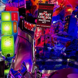 以下が含まれることがあります： ミュータントコントロールアンドレジストレーション法を支持する。ミュータントはこれ以上許さない！と書かれた看板のあるピンボールマシン。看板は、建物と赤と青の背景を持つ都市の風景の前にある。