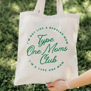 May include: Cream-colored tote bag with green text that reads "I'm not like a regular mom, I'm a Type One Mom, Type One Moms Club." The bag has two handles and is held in front of a green background.