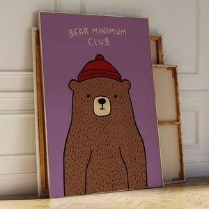 May include: A framed art print featuring a brown bear wearing a red knit cap. The bear is set against a purple background, with the words "BEAR MINIMUM CLUB" in white text at the top. The artwork is leaning against a wall.