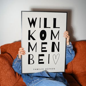 Könnte beinhalten: Ein rechteckiges gerahmtes Wandbild mit hellbeigem Hintergrund und fetter schwarzer Schrift, die WILLKOMMEN BEI lautet, gefolgt von einem handgezeichneten Herzumriss und dem Text FAMILIE HÜFKEN in einer kleineren, serifenlosen Schrift.