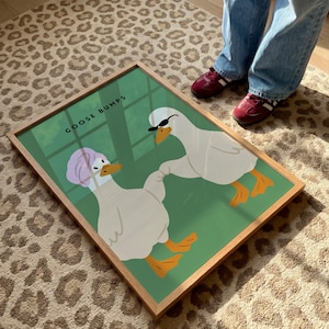 May include: A framed art print featuring two cartoon geese on a green background. One goose wears a pink head wrap, the other wears sunglasses. The words "GOOSE BUMPS" are printed at the top. The frame is made of light wood.