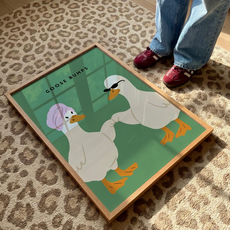 May include: A framed art print featuring two cartoon geese on a green background. One goose wears a pink head wrap, the other wears sunglasses. The words "GOOSE BUMPS" are printed at the top. The frame is made of light wood.