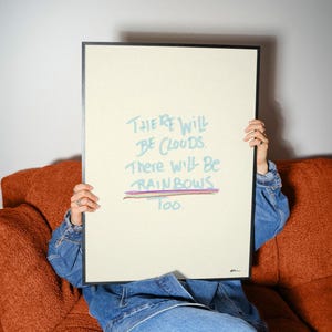 Könnte beinhalten: Ein rechteckiger gerahmter Druck mit hellcremefarbenem Hintergrund und einem dünnen schwarzen Rahmen. Der Text lautet "There will be clouds. There will be rainbows too." in Hellblau, mit einer mehrfarbigen horizontalen Linie unter dem Text.