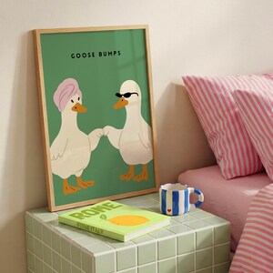 May include: A framed art print with the text "GOOSE BUMPS" features two cartoon ducks holding hands. One duck wears a pink turban, the other sunglasses. A book titled "ROME ITALY" and a blue and white striped mug are on a green tiled surface.