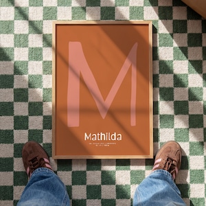 Könnte beinhalten: Ein gerahmter Kunstdruck mit dem Namen "Mathilda" in weißer Schrift unter einem großen, hellrosa Buchstaben "M" auf einem ziegelroten Hintergrund. Der Rahmen ist hellbraun. Der Druck liegt auf einem grün-weißen Schachbrettmuster-Teppich.