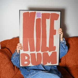 Könnte beinhalten: Ein gerahmtes Poster mit den Worten "NICE BUM" in großen, fetten, orangefarbenen Buchstaben. Das Poster hat einen hellbeigen Hintergrund und ein kleines lila Quadrat über dem Buchstaben "I". Die Person, die das Poster hält, trägt eine Jeansjacke und sitzt auf einem orangefarbenen Sofa.