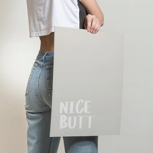 Modern wall art poster with a simple white background and light grey text reading “Nice Butt.” The design is minimalist, focusing on typography and negative space. Displayed in a lifestyle mockup, it complements Scandinavian and clean aesthetic interiors, making it ideal for stylish and understated home decor.