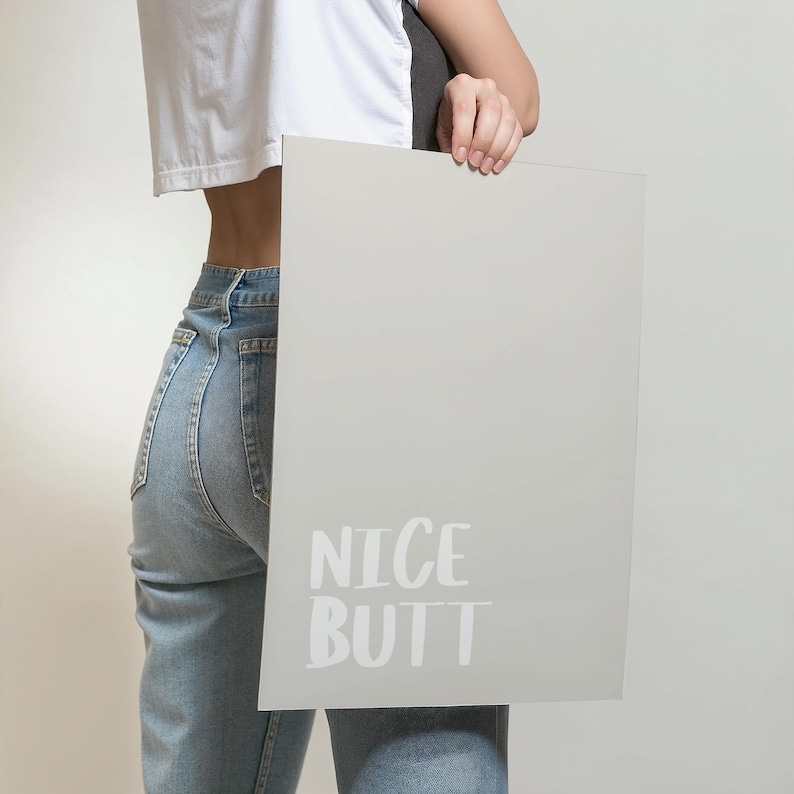 Modern wall art poster with a simple white background and light grey text reading “Nice Butt.” The design is minimalist, focusing on typography and negative space. Displayed in a lifestyle mockup, it complements Scandinavian and clean aesthetic interiors, making it ideal for stylish and understated home decor.