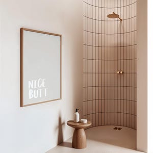 Minimalist white and light grey typography poster featuring the phrase “Nice Butt” in a clean modern font. The artwork is shown in a neutral setting, held by a person wearing casual clothing, emphasizing its contemporary and subtle humor style. Perfect for modern interiors, bathrooms, or bedrooms seeking a playful yet refined decorative element.