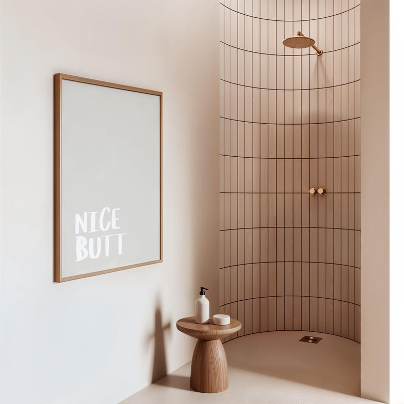 Minimalist white and light grey typography poster featuring the phrase “Nice Butt” in a clean modern font. The artwork is shown in a neutral setting, held by a person wearing casual clothing, emphasizing its contemporary and subtle humor style. Perfect for modern interiors, bathrooms, or bedrooms seeking a playful yet refined decorative element.