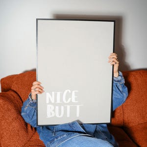 Minimalist white and light grey typography poster featuring the phrase “Nice Butt” in a clean modern font. The artwork is shown in a neutral setting, held by a person wearing casual clothing, emphasizing its contemporary and subtle humor style. Perfect for modern interiors, bathrooms, or bedrooms seeking a playful yet refined decorative element.