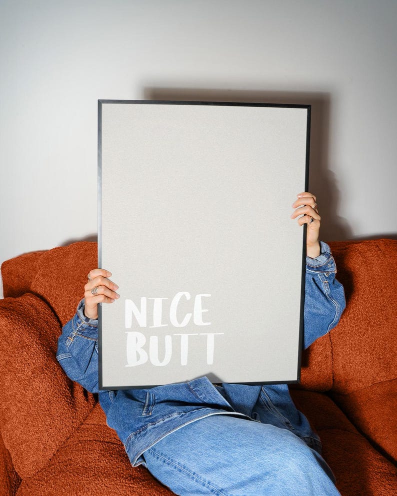 Minimalist white and light grey typography poster featuring the phrase “Nice Butt” in a clean modern font. The artwork is shown in a neutral setting, held by a person wearing casual clothing, emphasizing its contemporary and subtle humor style. Perfect for modern interiors, bathrooms, or bedrooms seeking a playful yet refined decorative element.
