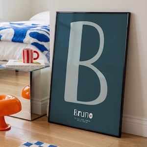 Könnte beinhalten: Gerahmter tealfarbener Druck mit einem großen weißen Buchstaben "B". Der Name "Bruno" ist darunter in Weiß gedruckt. Der Druck wird in einem Raum mit Bett und Beistelltisch angezeigt.