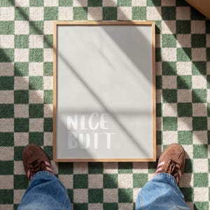 Minimalist white and light grey typography poster featuring the phrase “Nice Butt” in a clean modern font. The artwork is shown in a neutral setting, held by a person wearing casual clothing, emphasizing its contemporary and subtle humor style. Perfect for modern interiors, bathrooms, or bedrooms seeking a playful yet refined decorative element.