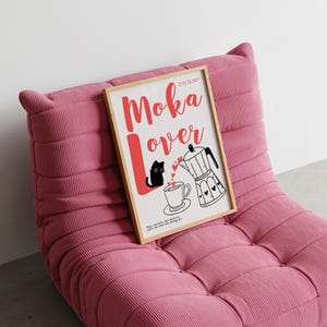May include: A framed print with the words "Moka Lover" in red script, featuring a black cat, coffee cup, and a moka pot illustration. The print is displayed on a pink corduroy lounge chair.