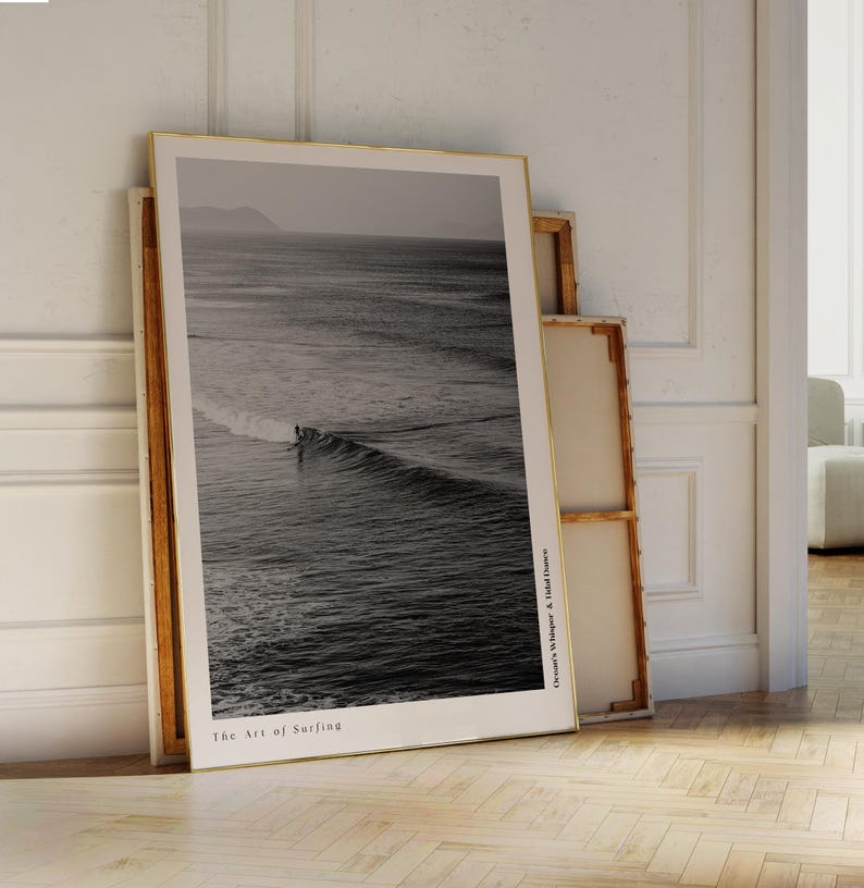 May include: A framed black and white print of a surfer riding a wave. The print has a gold frame and the text "The Art of Surfing" at the bottom. The print is leaning against a wall.