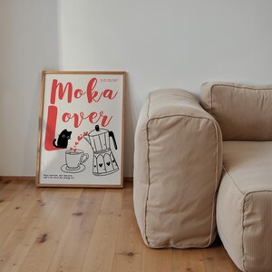 May include: A framed poster with the words "Moka Lover" in red script, featuring a black cat, a coffee cup, and a moka pot illustration. The poster is in a wooden frame and is set against a white wall, with a beige corduroy sofa in the background.