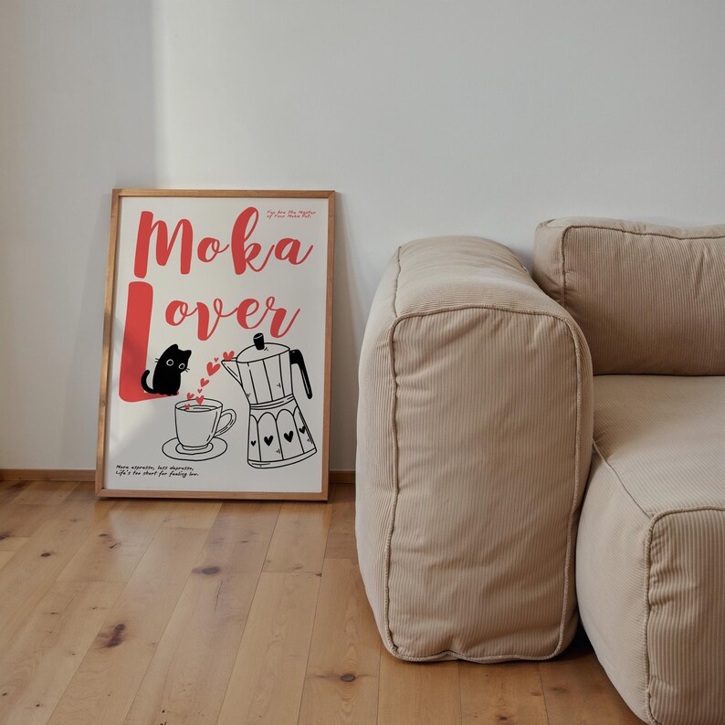 May include: A framed poster with the words "Moka Lover" in red script, featuring a black cat, a coffee cup, and a moka pot illustration. The poster is in a wooden frame and is set against a white wall, with a beige corduroy sofa in the background.