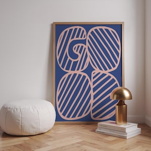 Könnte beinhalten: Ein gerahmter Kunstdruck mit dem Wort "GO" in rosa Streifen auf marineblauem Hintergrund. Eine goldene Lampe und ein weißer Hocker sind im Vordergrund zu sehen. Das Kunstwerk ist in einem Holzrahmen.