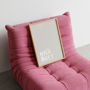 Modern wall art poster with a simple white background and light grey text reading “Nice Butt.” The design is minimalist, focusing on typography and negative space. Displayed in a lifestyle mockup, it complements Scandinavian and clean aesthetic interiors, making it ideal for stylish and understated home decor.
