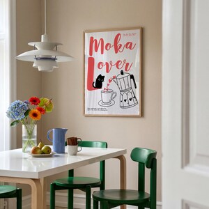 May include: A framed poster with the words "Moka Lover" in red script, featuring a black cat, coffee cup, and a moka pot. The poster is hanging on a beige wall above a white table with green chairs. A vase of flowers and fruit are on the table.