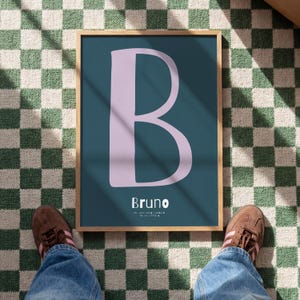 Op de afbeelding: Een ingelijste print met een grote, lichtpaarse letter "B" op een tealblauwe achtergrond. De naam "Bruno" staat eronder. De print zit in een licht houten frame, tegen een groen-wit geblokt patroon.