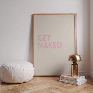 Könnte beinhalten: Ein gerahmter Druck mit den rosa Worten "GET NAKED". Eine goldfarbene Tischlampe steht auf Büchern. Links befindet sich ein weißer, runder Ottoman.