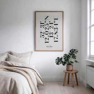 Peut inclure: Une affiche encadrée avec les coordonnées de Berlin en texte noir. L'affiche est dans un cadre clair et est accrochée à un mur blanc. Un lit avec une literie à rayures et un petit tabouret en bois avec une plante en pot sont au premier plan.